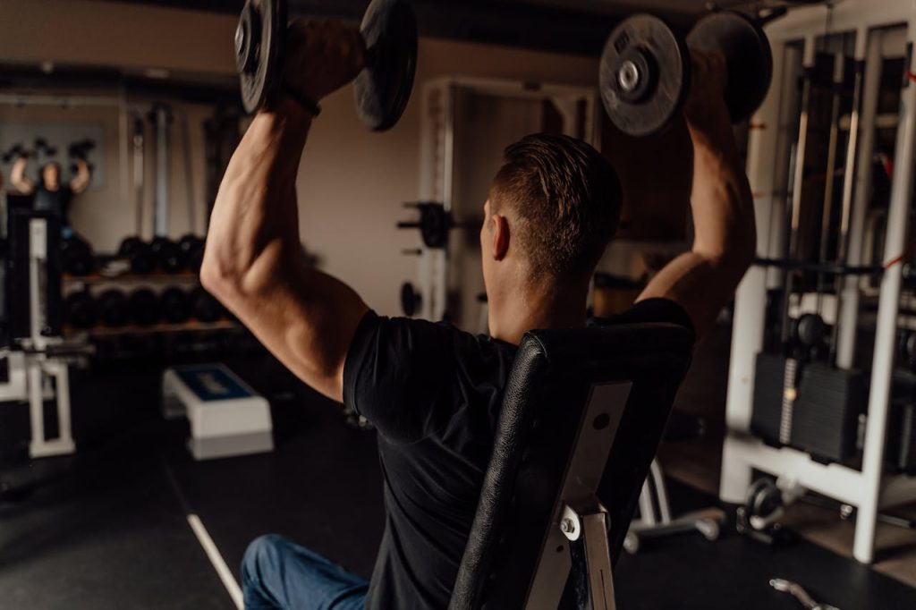 A muscular man seen from behind, wearing a black t-shirt, sitting on an adjustable bench and pressing two heavy dumbbells overhead in a gym setting