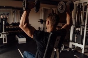 A muscular man seen from behind, wearing a black t-shirt, sitting on an adjustable bench and pressing two heavy dumbbells overhead in a gym setting